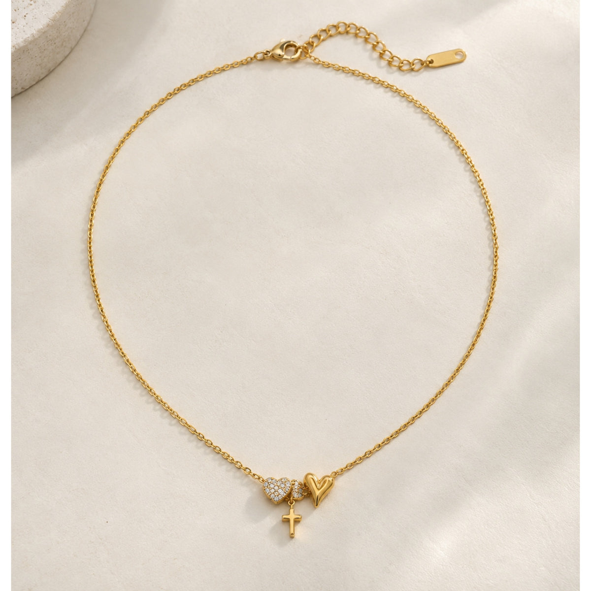 Collar de cadena fina en tono dorado con corazones lisos y decorados con zirconias, pequeña cruz colgante y extensión, sobre fondo beige.