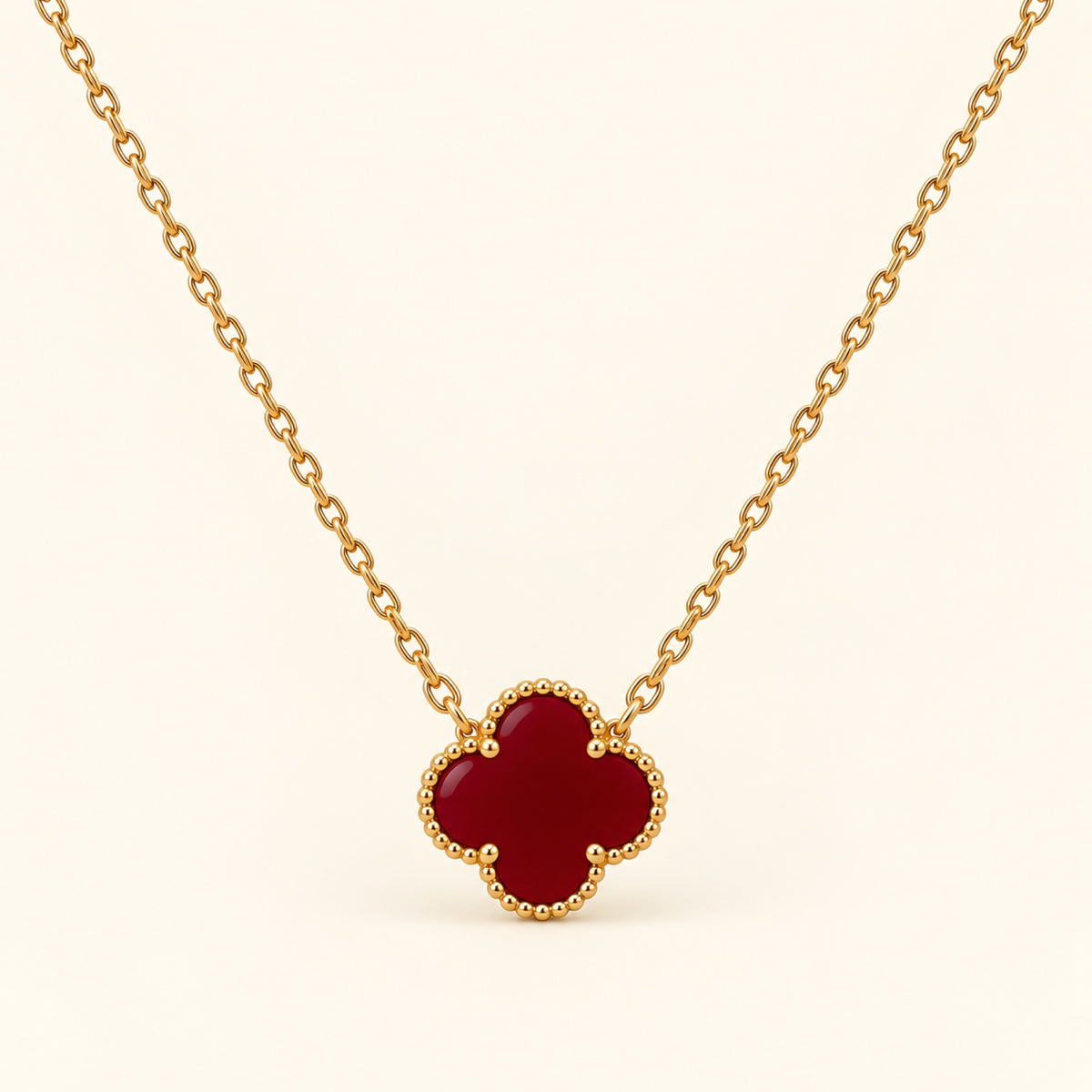 Collar de cadena dorada fina con trébol de cuatro hojas en rojo vino y borde de cuentas doradas sobre fondo marfil.