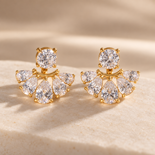 Par de aretes en tono dorado con zirconia redonda superior y abanico de cinco zirconias en forma de gota, sobre piedra beige.

