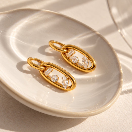 Par de aretes en tono dorado con argolla superior y marco ovalado colgante con zirconias rectangular, redonda y tipo marquesa, sobre fondo blanco.
