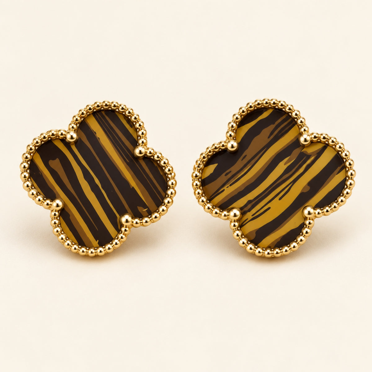 Par de aretes de trébol de cuatro hojas con franjas café y doradas y borde de pequeñas cuentas doradas sobre fondo marfil.