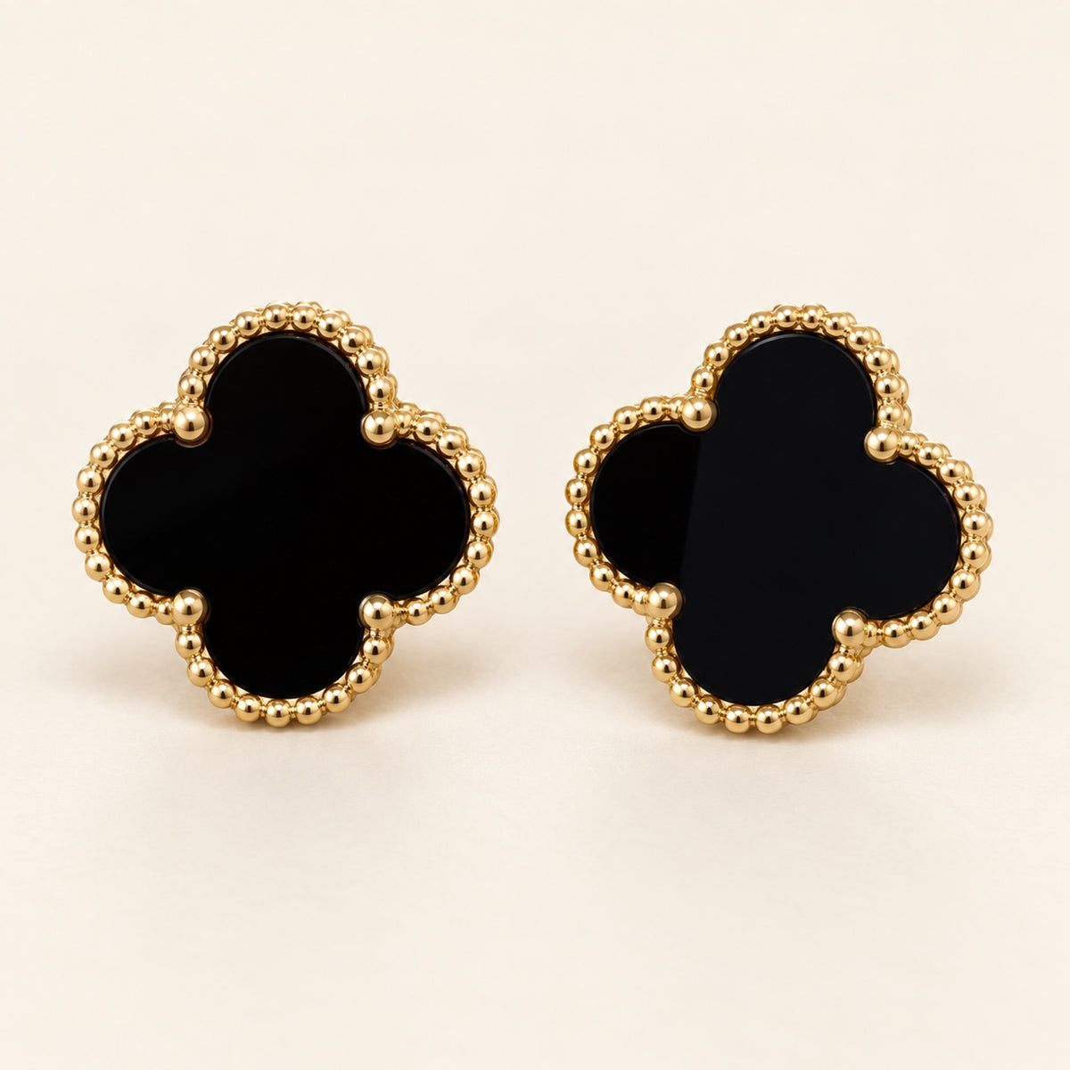 Par de aretes de trébol de cuatro hojas con centro negro y borde de cuentas doradas sobre fondo marfil.