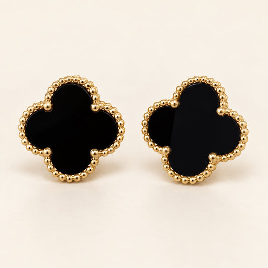 Par de aretes de trébol de cuatro hojas con centro negro y borde de cuentas doradas sobre fondo marfil.