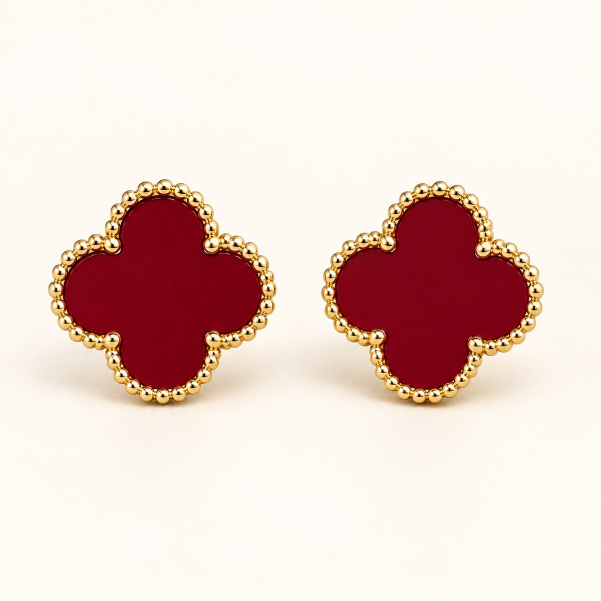 Par de aretes de trébol de cuatro hojas en rojo vino con borde de cuentas doradas sobre fondo marfil.