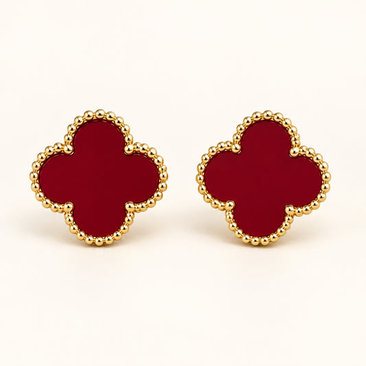 Par de aretes de trébol de cuatro hojas en rojo vino con borde de cuentas doradas sobre fondo marfil.