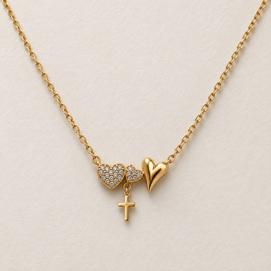Collar dorado con tres corazones, dos con zirconias y uno liso alargado, y una pequeña cruz colgante sobre fondo marfil.