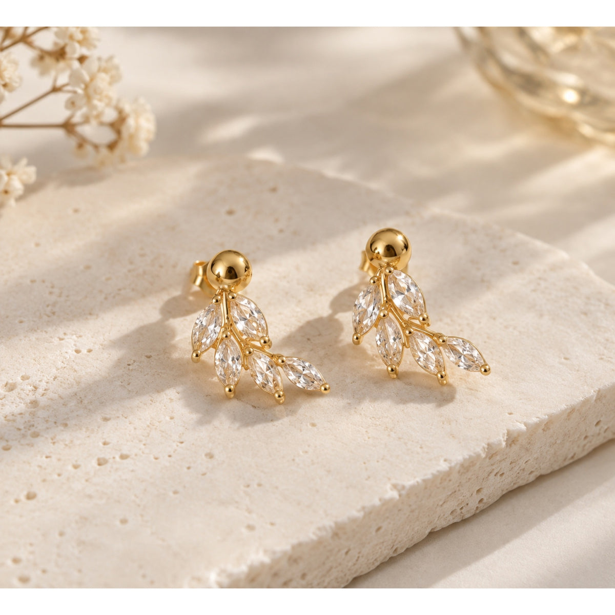 Par de aretes en tono dorado con bolita superior y cinco zirconias en forma de hoja dispuestas como una rama, sobre piedra beige.
