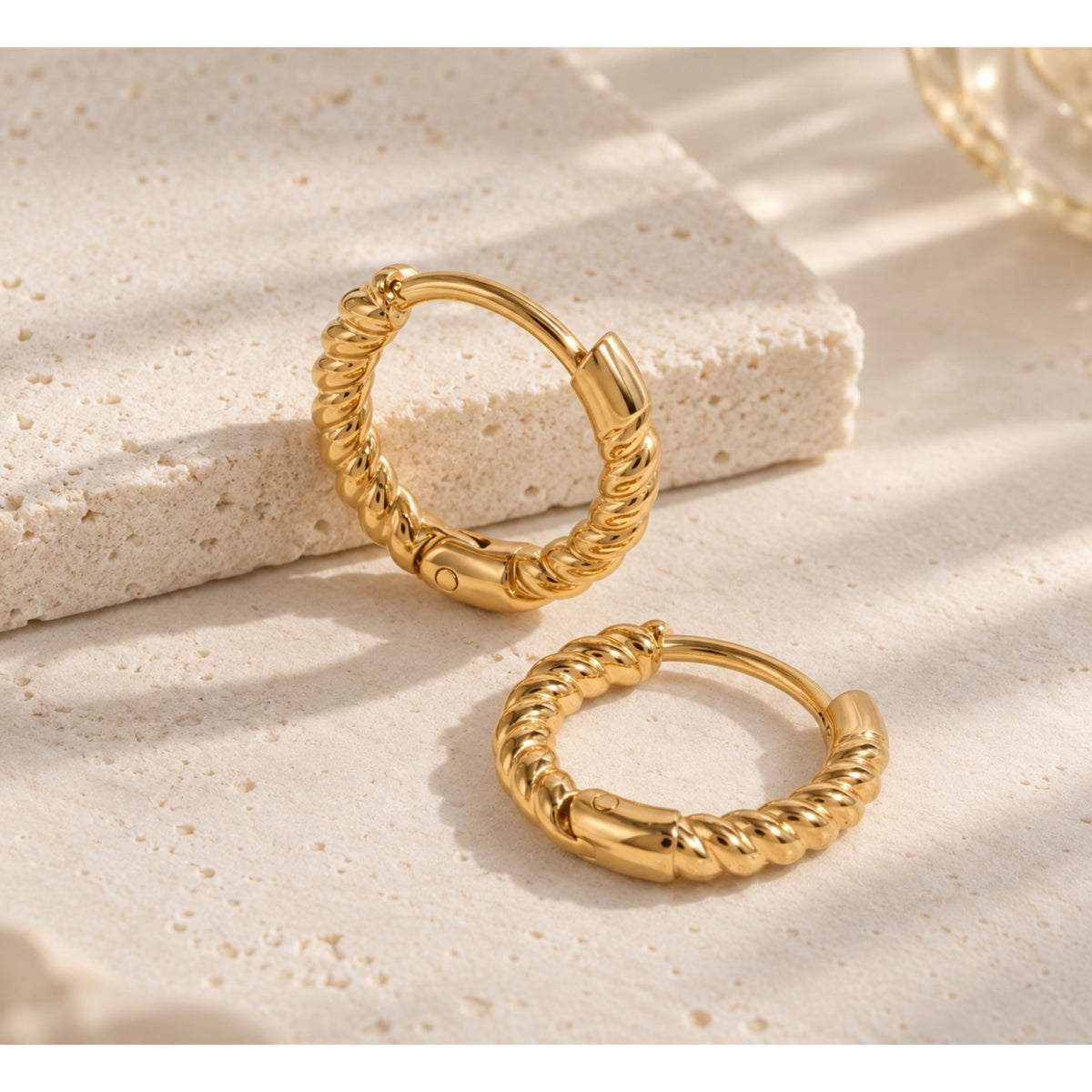 Par de argollas en tono dorado con textura torcida tipo cuerda y cierre articulado, sobre una superficie de piedra beige.