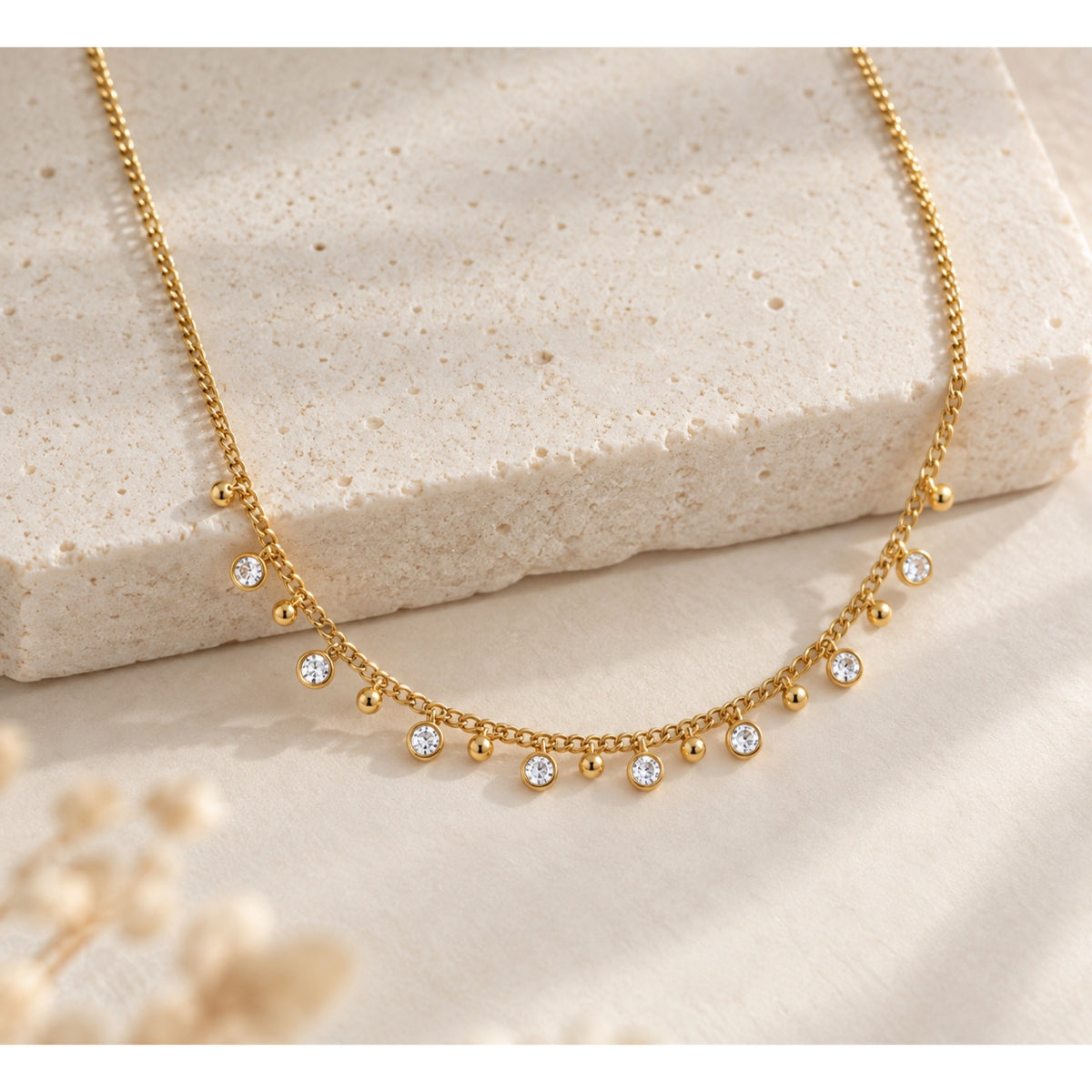 Collar en tono dorado con zirconias redondas colgantes intercaladas con pequeñas bolitas lisas, sobre una superficie de piedra beige.