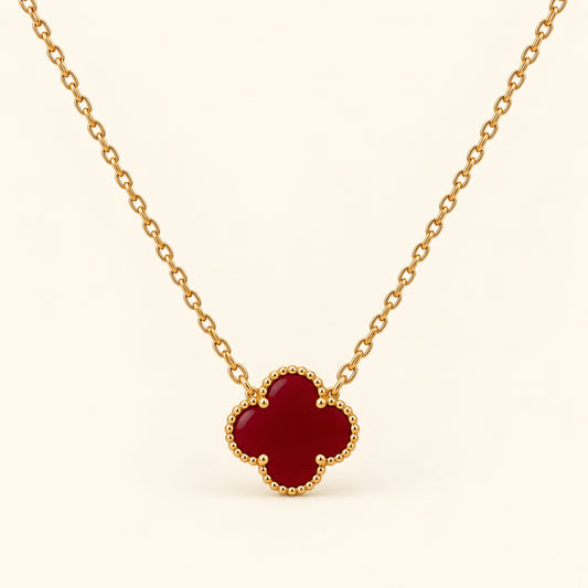 Collar de cadena dorada fina con trébol de cuatro hojas en rojo vino y borde de cuentas doradas sobre fondo marfil.